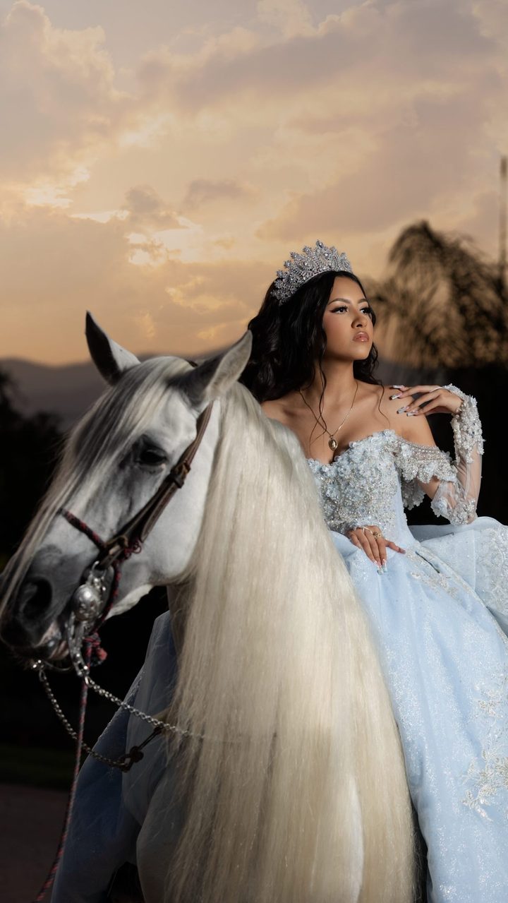 Quinceañera in a powder-blue gown riding a white horse at sunset