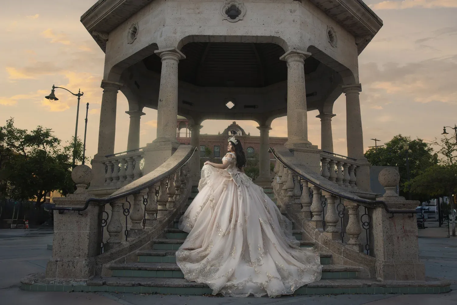 Quinceañera in an ivory gown on the steps of a stone gazebo at sunset