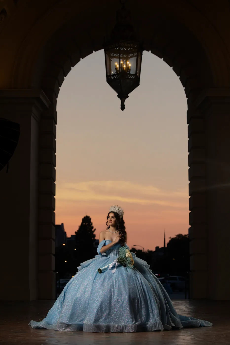 Quinceañera in a pale blue gown and crown seated beneath a stone arch at sunset