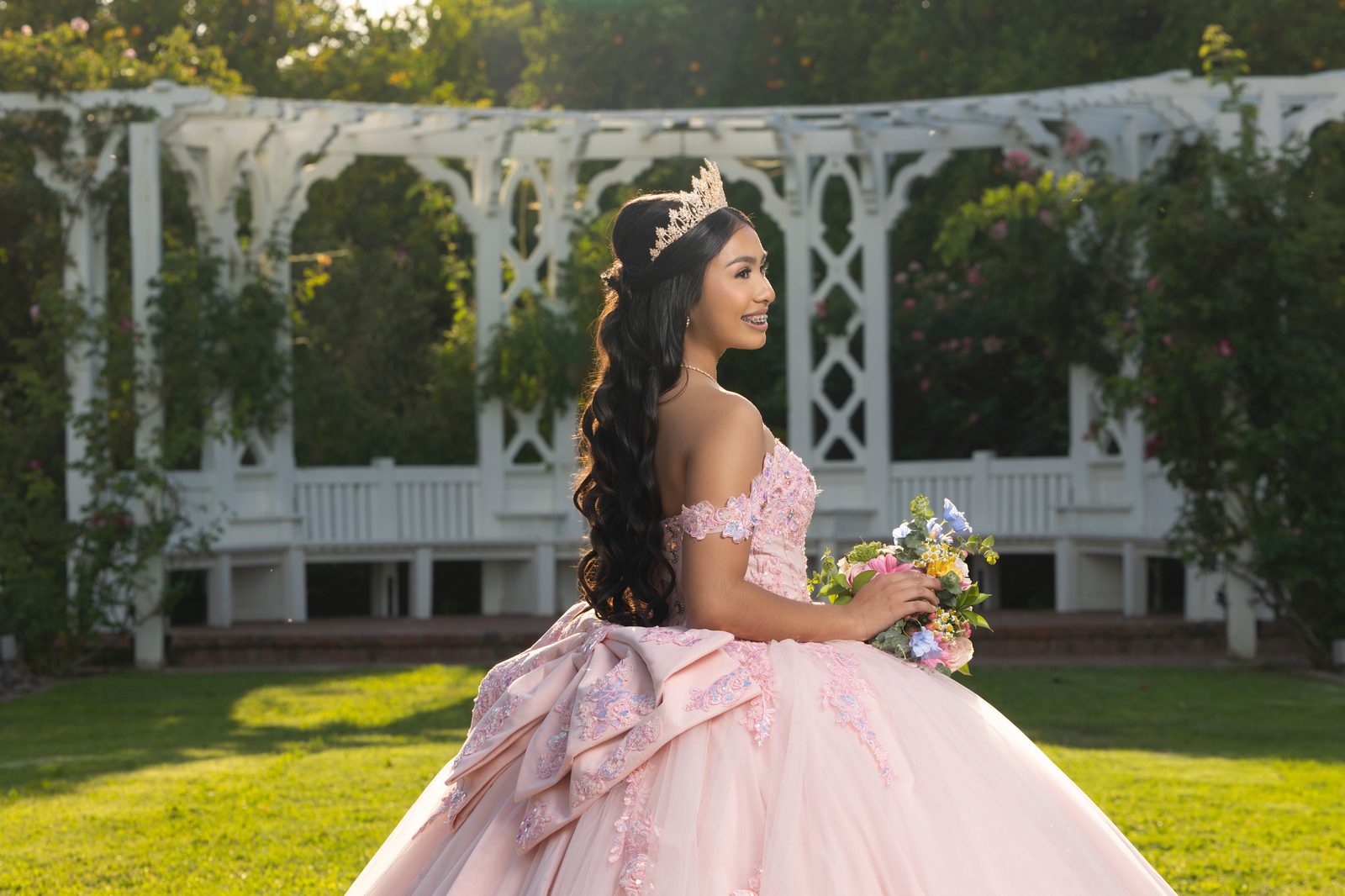 A blush gown with a satin bow and a bouquet in front of a white garden pergola