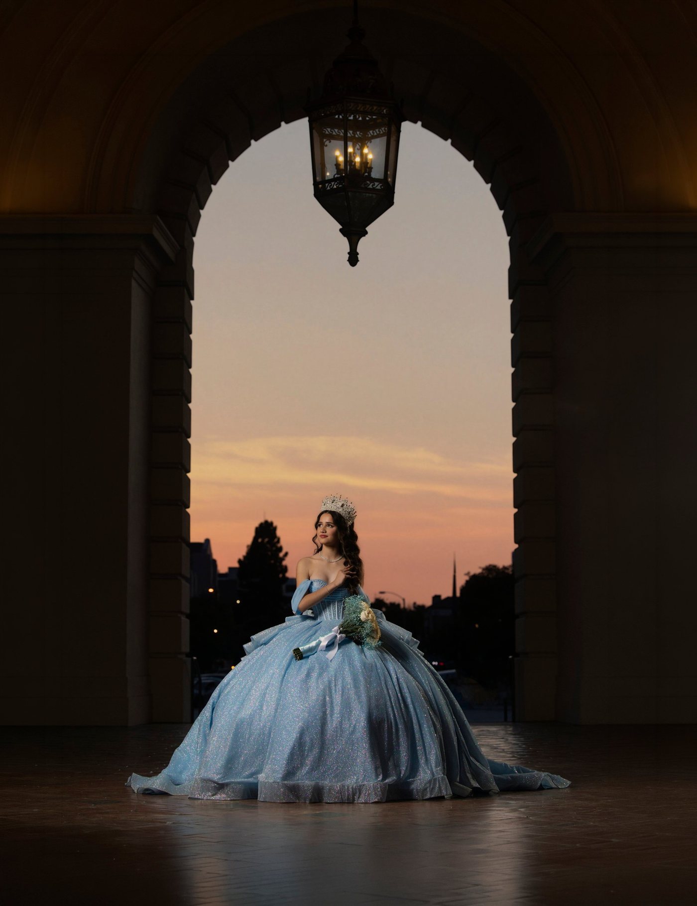 Quinceañera in a blue gown under the lantern-lit arch of Pasadena City Hall at sunset